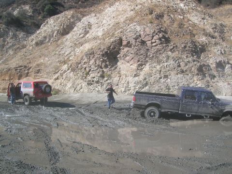 1985 Ford Bronco - Two vehicles are stuck in a muddy area, with people trying to free them.