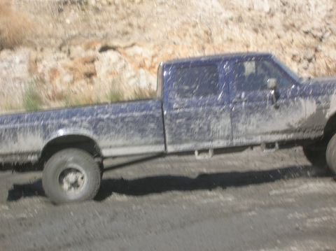 1985 Ford Bronco - A muddy blue pickup truck navigates a dirt road with a rocky hillside in the background.