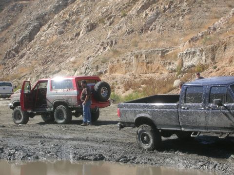 1985 Ford Bronco - A red 1985 Ford Bronco is parked next to a muddy blue pickup truck on a dirt road with a rocky hillside in the background.