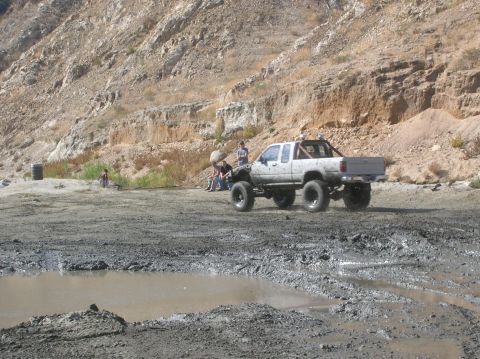 1985 Ford Bronco - Off-road pickup truck navigates a muddy terrain with onlookers present.