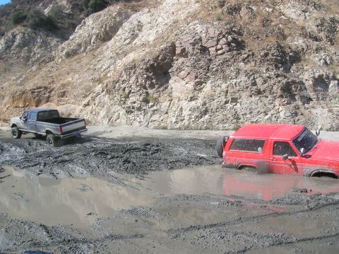 1985 Ford Bronco - Two off-road vehicles are deeply mired in mud on a treacherous dirt path.