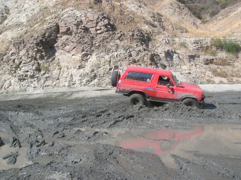 1985 Ford Bronco - A red 1985 Ford Bronco navigates a muddy off-road trail, with its rear spare tire prominent.