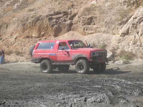 1985 Ford Bronco - Muddy red Ford Bronco navigating rough terrain. Grille design suggests an early 1980s model.