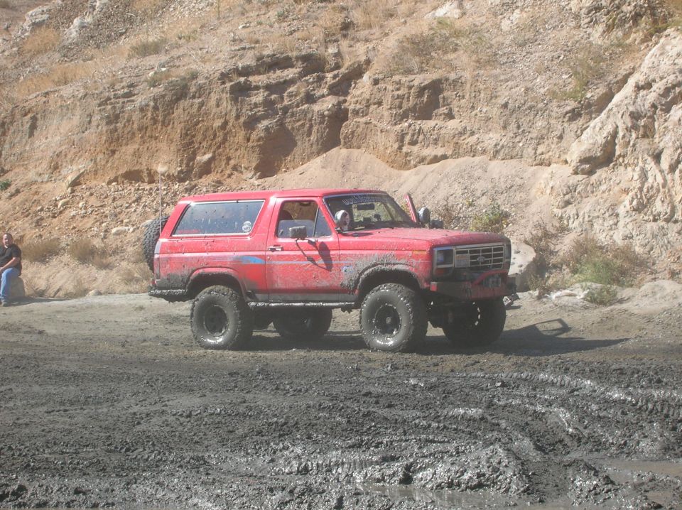 Red Ford Bronco with off-road tires stuck in mud, side view. Grille indicates early 1980s model