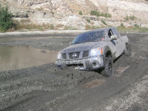 1985 Ford Bronco - A Nissan pickup truck is stuck in thick mud, with its front end submerged in a rut.