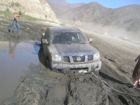 1985 Ford Bronco - A Nissan Titan truck is stuck in thick mud, with a person standing nearby and another vehicle in the background.