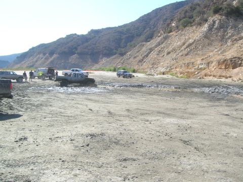 1985 Ford Bronco - Multiple trucks are seen navigating a muddy off-road track in a dry, hilly landscape.
