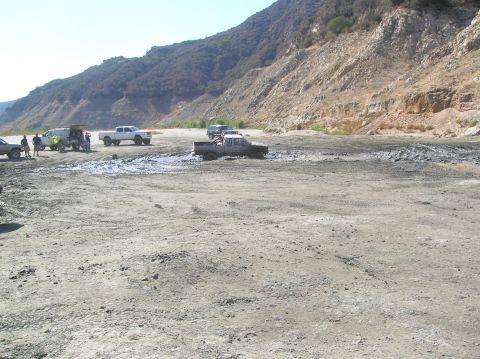 1985 Ford Bronco - Multiple trucks are stuck in mud near a rocky hillside.