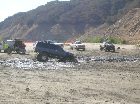 1985 Ford Bronco - Several trucks are in a muddy area with a hillside in the background.