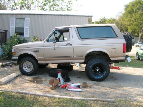 1996 Ford Bronco - Driver-side view of a tan Ford Bronco mid-way through a tire and suspension job in a gravel driveway, with a large Pro Comp Mud Terrain tir…