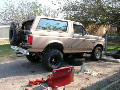 1996 Ford Bronco - Driver-side rear three-quarter view of a tan Ford Bronco with hardtop mid wheel-and-tire swap in a gravel driveway, wearing a new black-ste…