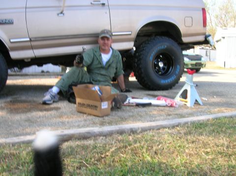 1996 Ford Bronco - A man in green coveralls and a ball cap sits on the gravel beside a tan Ford Bronco during a suspension install, with a Rough Country parts…