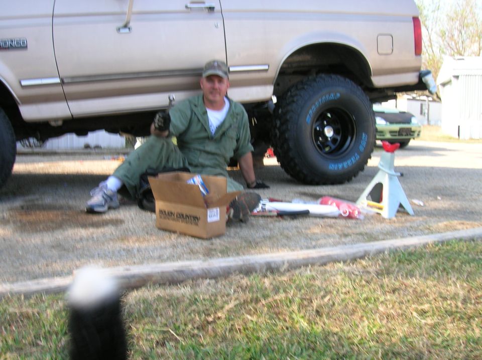 Man in coveralls installing Rough Country suspension parts on a tan Ford Bronco