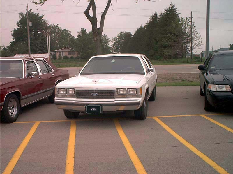 White Ford Crown Victoria parked in a lot, front view. The car has a chrome grille and rectangular headlights