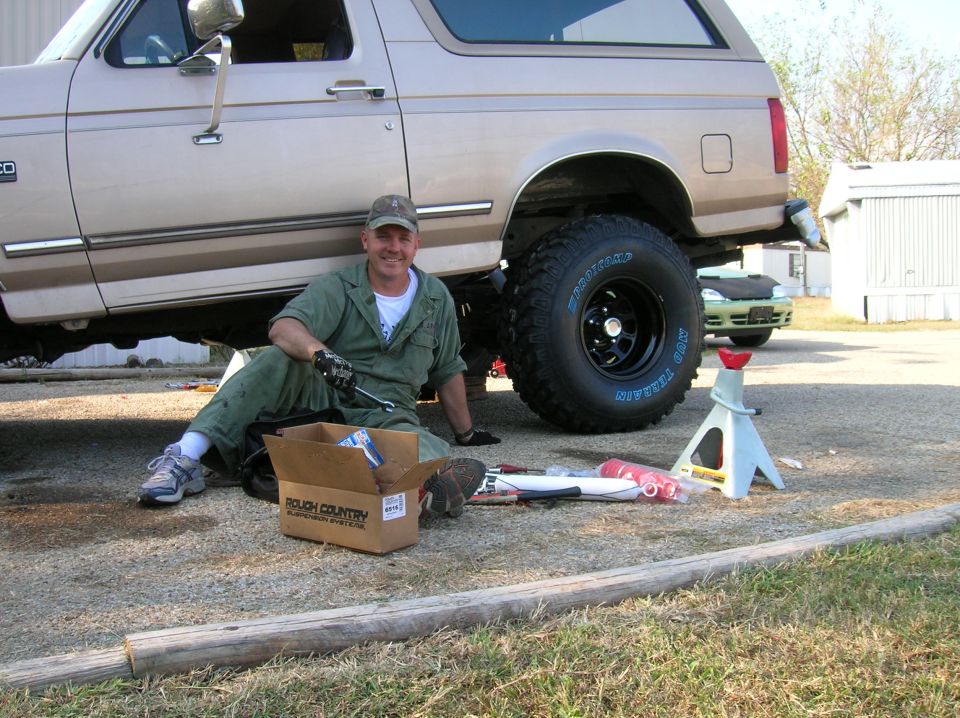 Tan Ford Bronco with lifted suspension and large tires, a man in green overalls sits nearby with tools