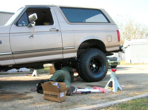 1996 Ford Bronco - Mechanic works on the underside of a lifted 1996 Ford Bronco, with a Rough Country suspension box nearby.