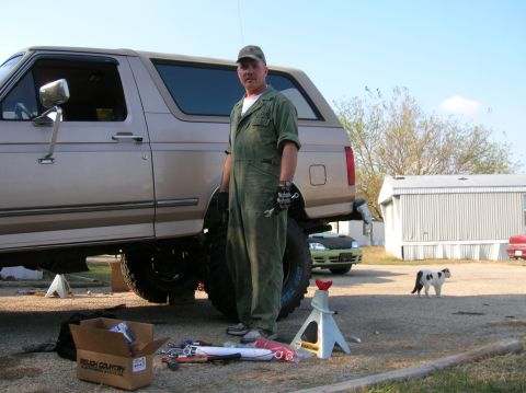 1996 Ford Bronco - A man in green coveralls and a ball cap stands in a gravel driveway beside a tan two-door SUV mid lift install, holding a wrench and a 'Mec…