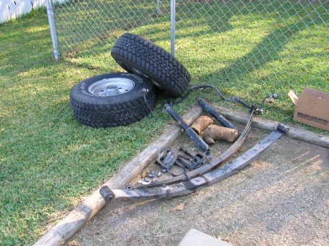 1996 Ford Bronco - Take-off suspension parts laid out on grass beside a chain-link fence: two rear leaf-spring packs, a sway bar with end links, four black sh…