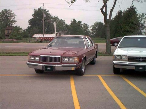 2000 Ford Crown Victoria - A maroon sedan is parked in a lot next to a white sedan, with trees and buildings in the background.