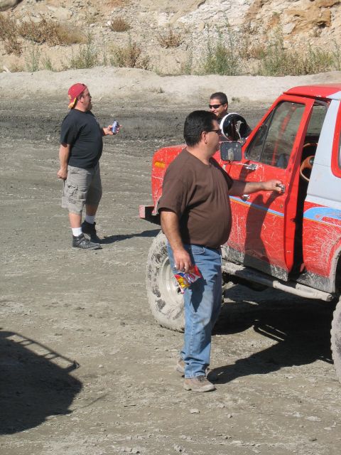 1987 Ford Bronco - A man opens the door of a red off-road vehicle while another man stands nearby holding a can in a dusty outdoor area.