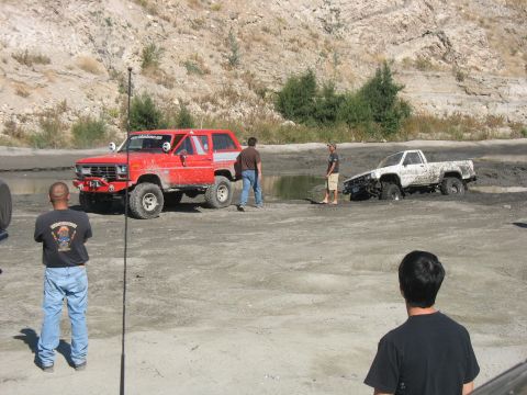 1987 Ford Bronco - Two off-road vehicles, a red SUV and a white pickup, are stuck in a muddy area with onlookers observing.
