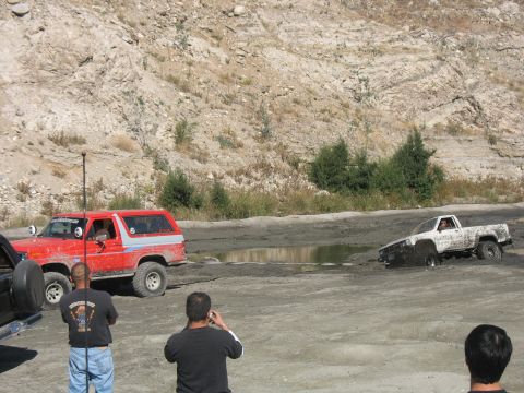 1987 Ford Bronco - Two off-road vehicles navigate a challenging muddy terrain, with one truck appearing to be stuck.