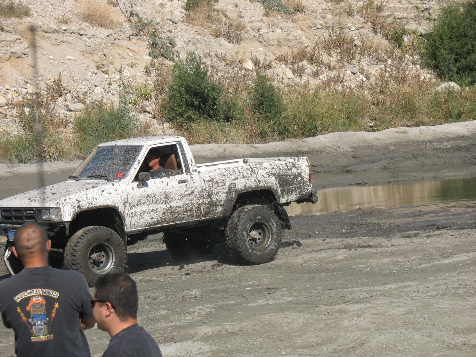 Mud-covered white pickup truck with large tires driving through a muddy puddle. Two people stand in the foreground