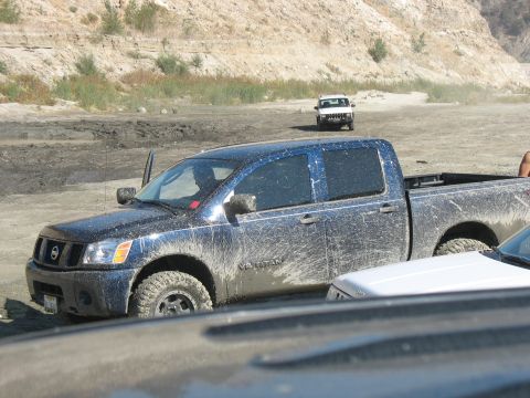 1987 Ford Bronco - A muddy Nissan Titan truck is parked off-road with a white SUV in the distance.
