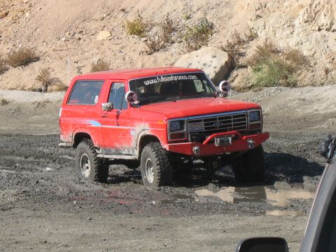1987 Ford Bronco - A red Ford Bronco navigates a muddy off-road course, kicking up water.