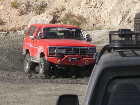 1987 Ford Bronco - A red Ford Bronco navigates a muddy off-road course, its front grille and body style indicating an early 1980s model year.