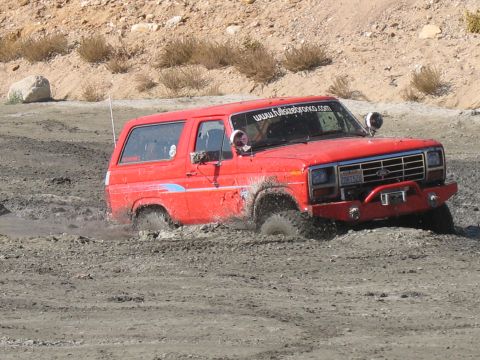 1987 Ford Bronco - This red Ford Bronco is stuck in the mud, with visible website and license plate information.
