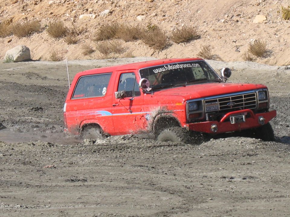 Red Ford Bronco with blue stripe stuck in mud, front 3/4 view. Visible text: www.fullsizebronco.com and license plate 2DB7524