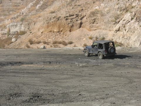 1987 Ford Bronco - A dark blue Jeep Wrangler is seen from the rear, parked on a dusty, uneven terrain with a rocky hillside in the background.