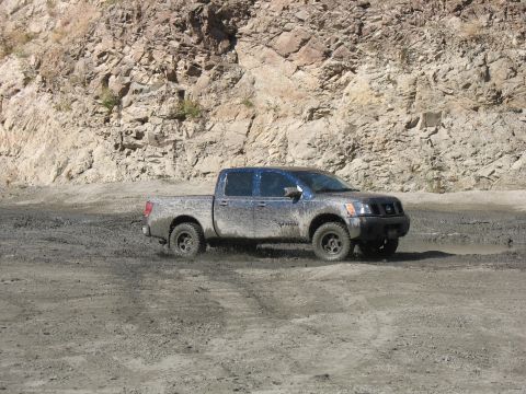 1987 Ford Bronco - Nissan Titan pickup truck navigating a muddy terrain with a rocky cliff face behind it.