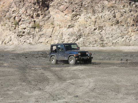 1987 Ford Bronco - A blue Jeep Wrangler navigates a muddy terrain with a rocky cliff face in the background.
