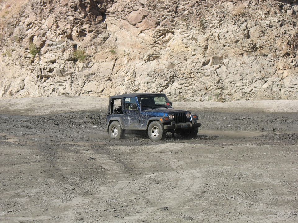 Blue Jeep Wrangler driving through mud near a rocky hill. Supermotors.net logo visible