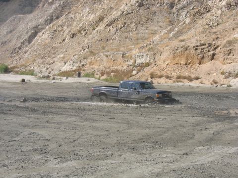 1987 Ford Bronco - A Ford F-Series truck navigates a muddy terrain with a rocky hillside in the background.