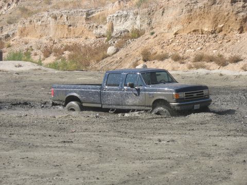 1987 Ford Bronco - A muddy 1987 Ford F-Series pickup truck is stuck in a dirt area, with its tires submerged in mud.