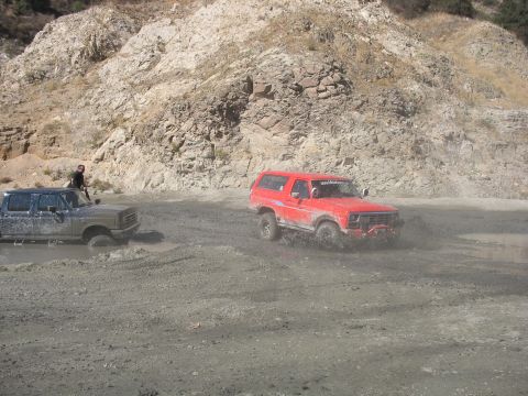 1987 Ford Bronco - Two off-road vehicles navigating a muddy terrain with a rocky hillside in the background.