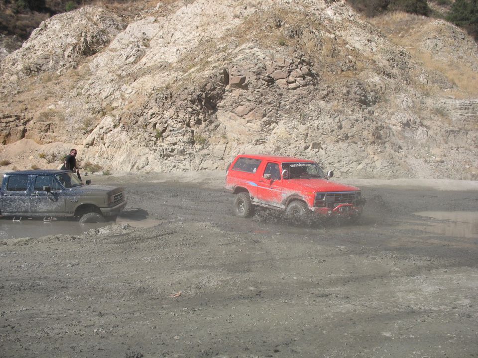 1987 Ford Bronco splashing through mud, another truck stuck behind it