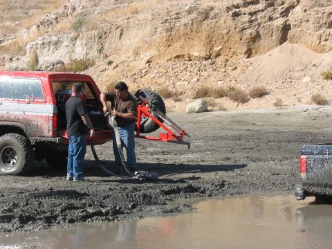1987 Ford Bronco - Two men work on a winch attached to a red SUV stuck in mud, with another vehicle visible in the background.