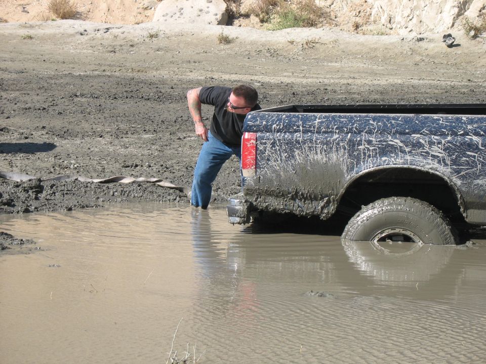 Rear view of a muddy 1987 Ford Bronco stuck in water, with a person checking it
