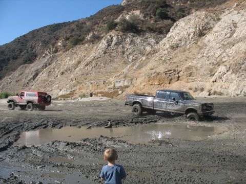 1987 Ford Bronco - A child watches as two off-road vehicles traverse a challenging muddy landscape.