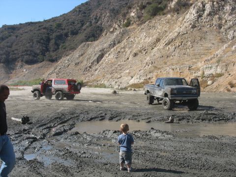 1987 Ford Bronco - Two lifted trucks navigate a muddy terrain under a clear sky, with a child observing the scene.