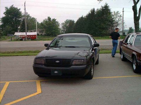 2000 Ford Crown Victoria - Front view of a black Ford Crown Victoria sedan parked in a lot with yellow-striped pavement, showing blacked-out grille surround, unpainte…