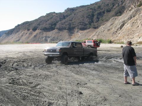 1987 Ford Bronco - A lifted Ford F-Series truck navigates muddy terrain with a red SUV in the background.