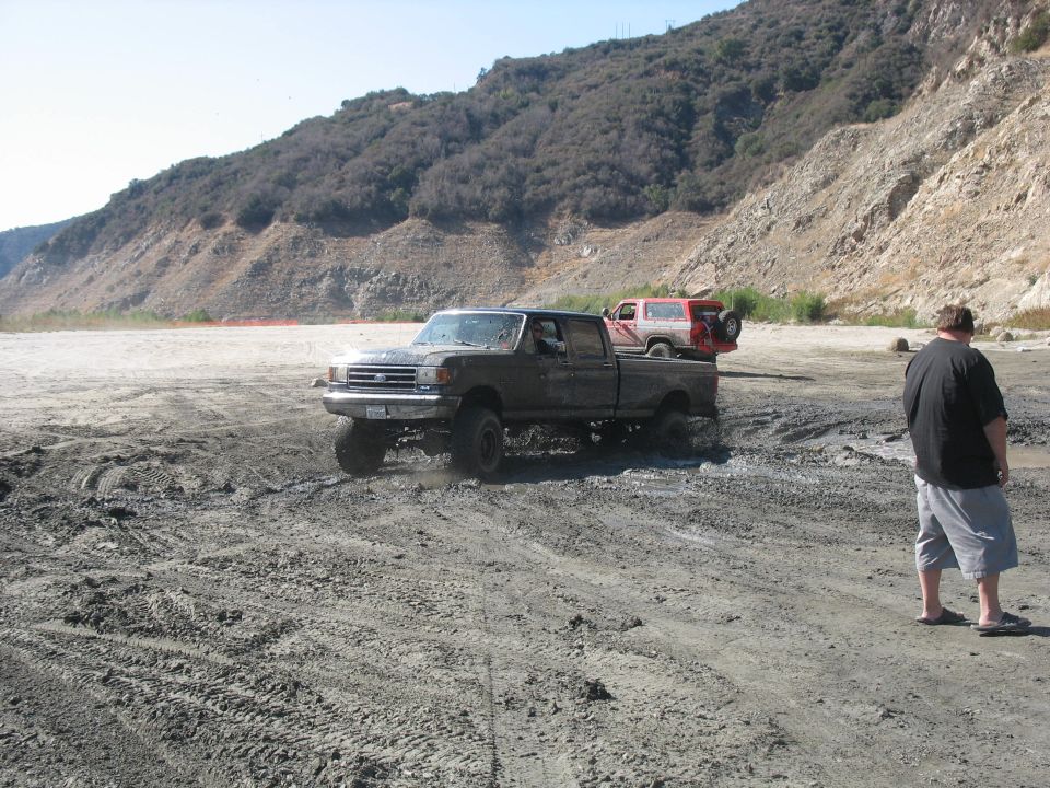 Dark gray Ford F-Series truck with lifted suspension in mud; red SUV behind it