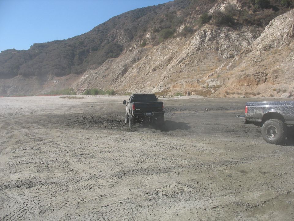 Two pickup trucks driving in a dusty, muddy area near a mountain