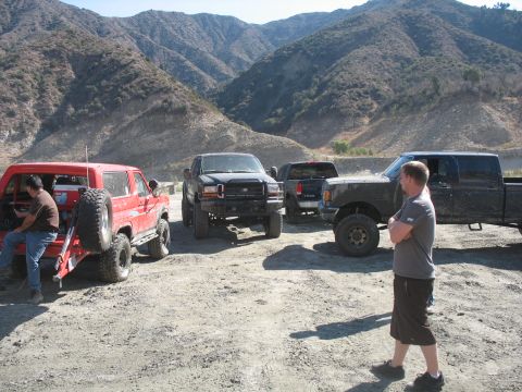 1987 Ford Bronco - Several off-road vehicles are parked in a dusty, mountainous area, with a man standing in the foreground.