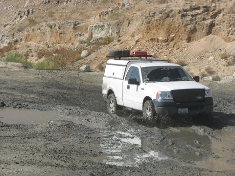 1987 Ford Bronco - A white pickup truck navigates a muddy terrain, its tires splashing through the mire.